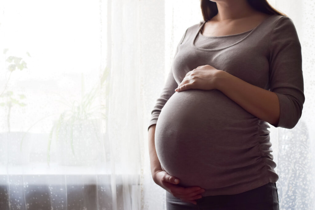 Pregnant woman with big belly at window. Maternity prenatal care and woman pregnancy concept. Young pregnant woman holds her hands on her swollen belly. Love concept. Horizontal with copy space.