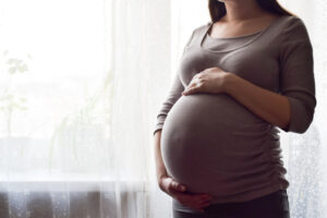 Pregnant woman with big belly at window. Maternity prenatal care and woman pregnancy concept. Young pregnant woman holds her hands on her swollen belly. Love concept. Horizontal with copy space.